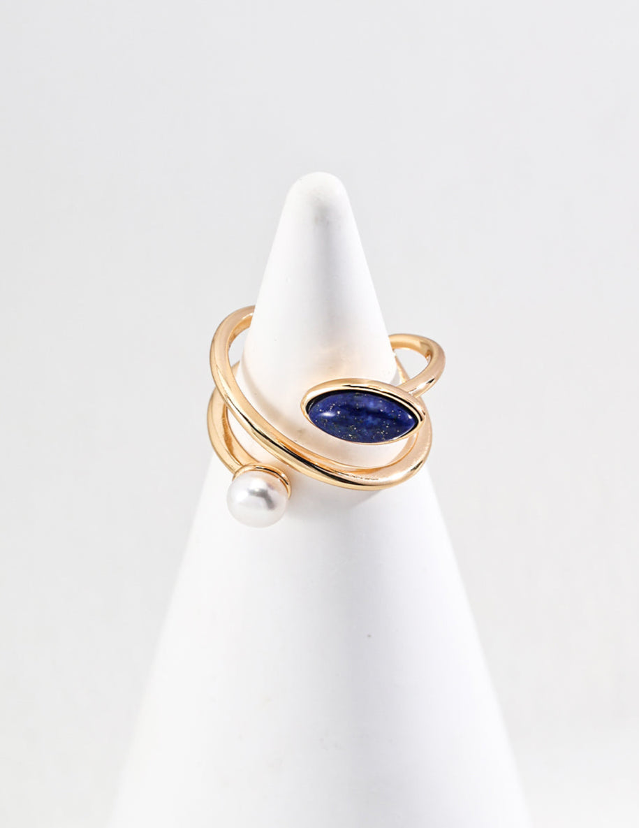 Gold ring with a blue gemstone and pearl on a white stand against a light gray background