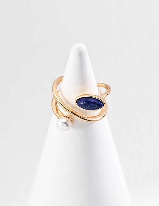 Gold ring with a blue gemstone and pearl on a white stand against a light gray background